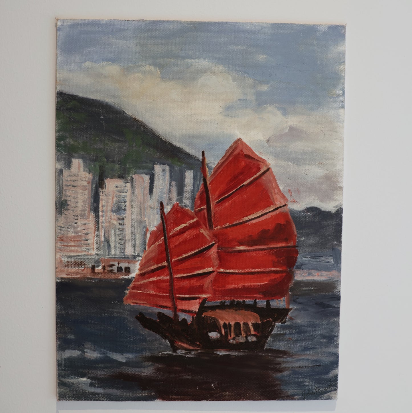 A painting of a traditional junk boat with vibrant red sails on a calm harbor, skyscrapers and a green hillside in the background.