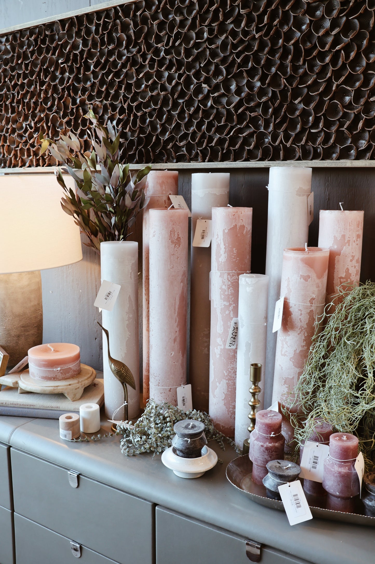 A decorative display of textured, cylindrical candles in soft pink, white, and purple hues, arranged with greenery, a lamp, and metallic accents.