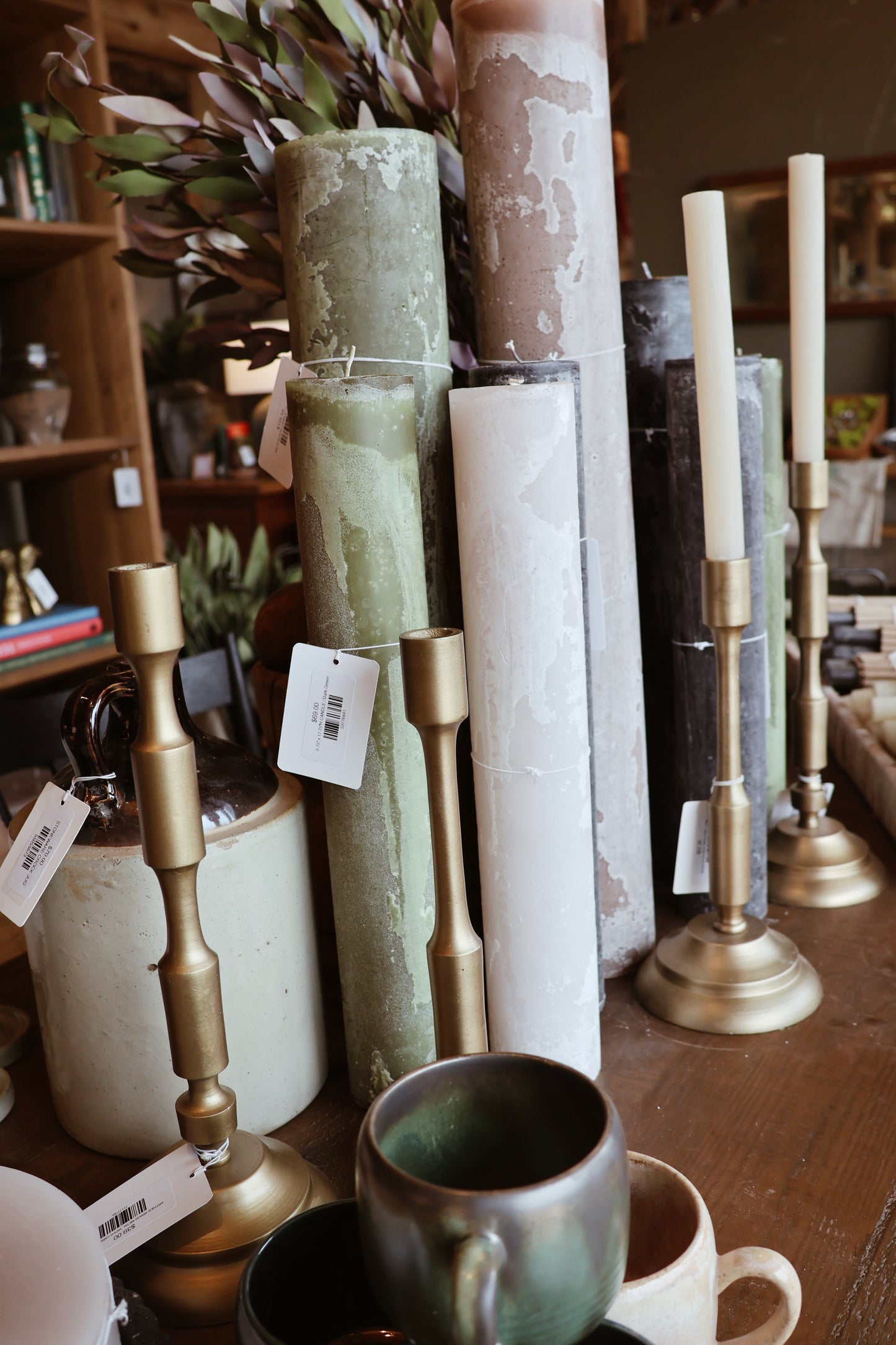 A cozy display of rustic pillar candles in earthy tones, paired with brass candlesticks, ceramic mugs, and decor items on a wooden table.