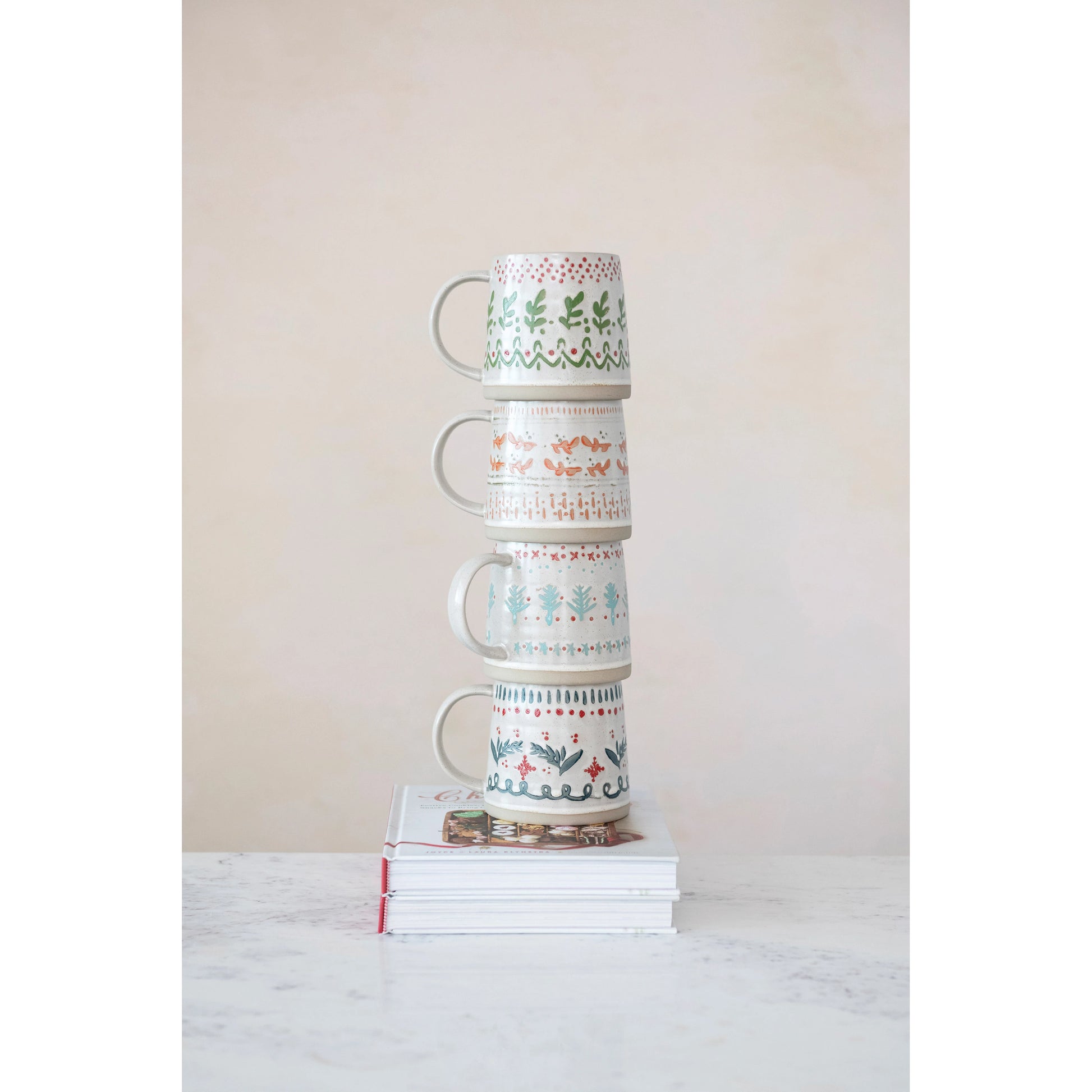 four ceramic mugs with festive, hand-painted designs featuring plants, patterns, and holiday-themed colors on a beige base.